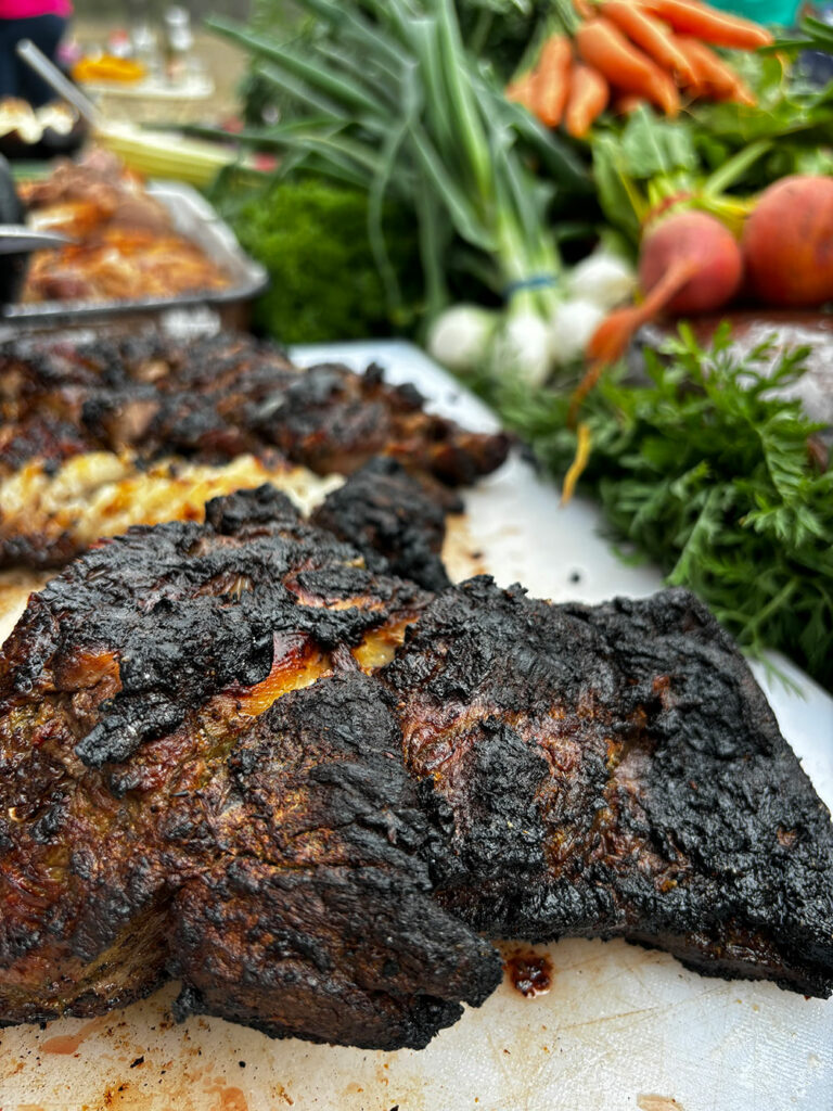A slab of woodfired meat sits in the foreground, surrounded by fresh farm produce. 